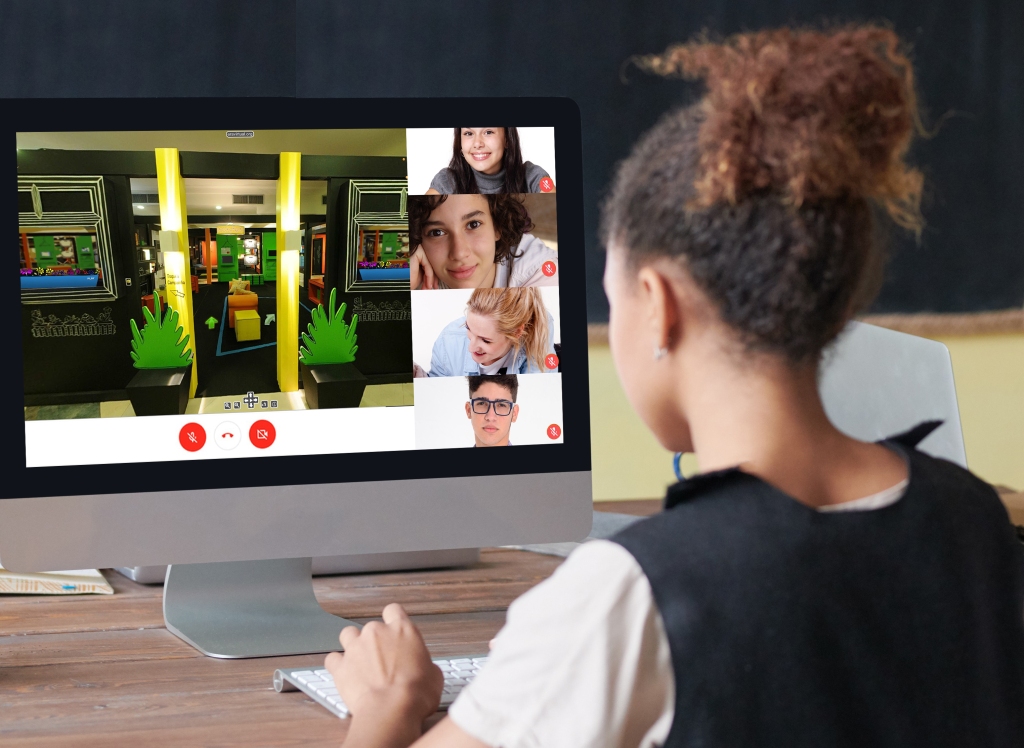 Fotografia de uma menina de costas, em frente ao computador, participando da visita virtual à exposição "Cadê a Química?", com seus quatro colegas de turma, que aparecem na tela em um chat online.