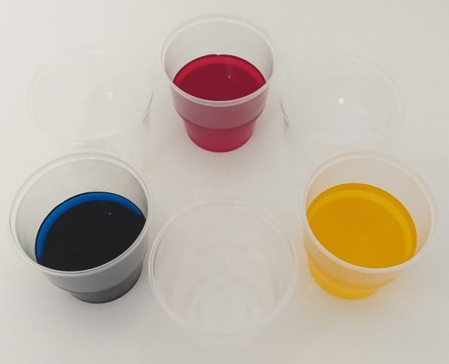 Fotografia de seis copos transparentes organizados em círculo. Três deles contêm água colorida por corantes azul, amarelo e vermelho e os outros três estão vazios.