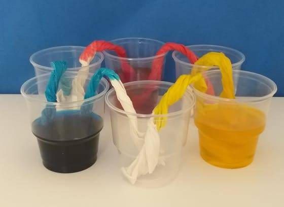 Fotografia de seis copos transparentes organizados em círculo com pontes/canudos de papel conectando eles. Três deles contêm água colorida por corantes azul, amarelo e vermelho e os outros três estão vazios. 