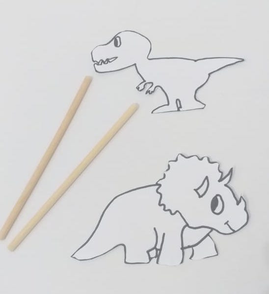 Imagem contendo o recorte de dois dinossauros e papel. Ao lado dos dinossauros, há dois palitinhos de madeira.