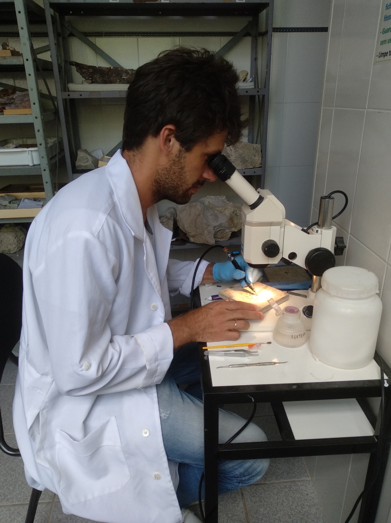 Um homem cientista, de jaleco branco, em uma mesa a observar através de uma lupa de laboratório fragmentos de fósseis.