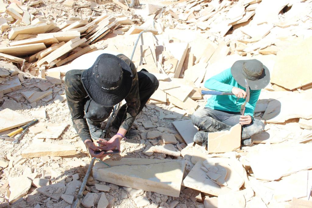 Duas pessoas sentadas sob um deposito de placas de fósseis a céu aberto, ambas com equipamentos de extração de fósseis em ação.