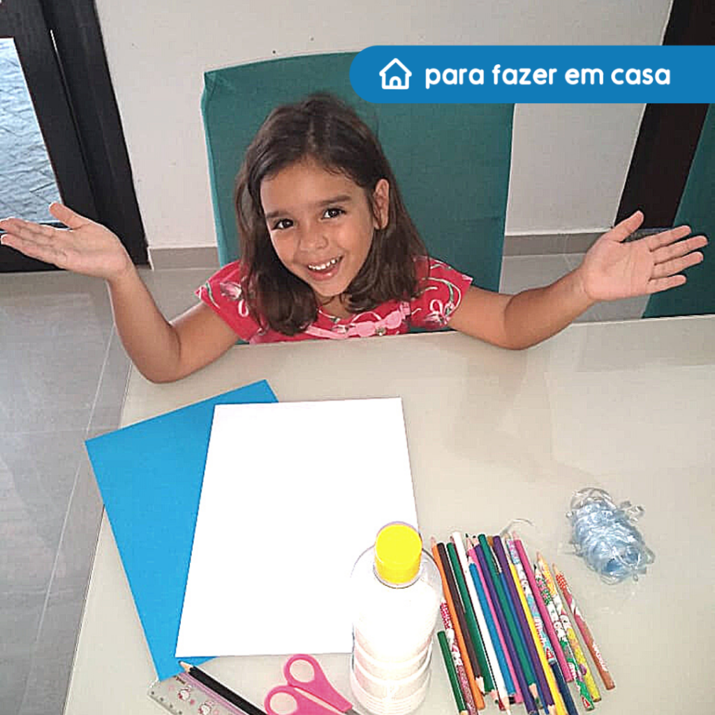 Fotografia de uma criança sentada com expressão de alegria e braços apoiados sobre uma mesa com folhas brancas e azuis, cola, tesoura, lápis de escrever, régua e lápis de cores.