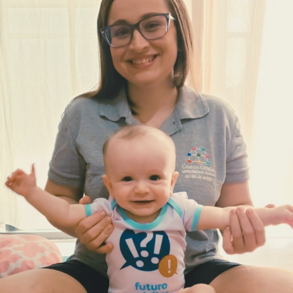 Fotografia de uma mãe branca, de cabelos castanhos, vestindo a camisa da Casa da Ciência, com um bebê branco sorridente, vestindo um body de futuro “minitor” da Casa da Ciência.