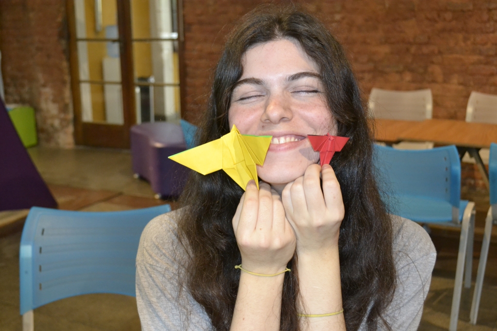 Fotografia de uma mediadora na Casa da Ciência, sorrindo com os olhos fechados e segurando duas borboletas de origami, uma em cada mão.
